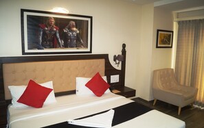 A hotel room with a bed, two red pillows, a headboard, a nightstand, a lamp, a chair, and a framed picture on the wall. - Phoenix Hotel (Bengaluru)
