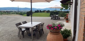 An outdoor patio with a table and chairs, a large umbrella, and a view of rolling hills and wind turbines in the background. - Old Parish House   (Orciano Pisano)