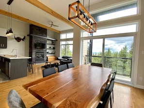 A modern kitchen with a stone fireplace, a dining area with a large wooden table, and large windows offering a view of the outdoors. - Chalet La Watch (Petite-Rivière-Saint-François)