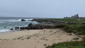 Ein Strand mit Felsformationen und einem historischen Gebäude im Hintergrund.