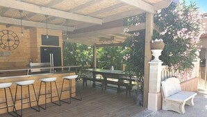 Un patio exterior con terraza de madera, una zona de bar con taburetes, una mesa de comedor y un banco.