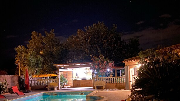 Un área de piscina exterior iluminada de noche, con dos sillones rojos, una estructura pequeña iluminada y vegetación circundante.