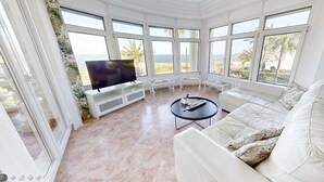 A spacious living room with a large white sofa, a flat-screen TV, and a coffee table. The room has large windows offering a view of the beach and palm trees. - Villa Frente al mar con Vistas, Terraza, Barbacoa y Piscina con spa (Bahía de Jávea)