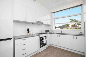 A modern kitchen with white cabinetry, a gas stove, a microwave, and a large window with a view of palm trees. - Domi Rentals - The Meadow Gardens (Saint Kilda East)