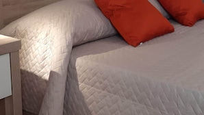 Quarto com cabeceira de madeira, cama com cobertura acolchoada, duas almofadas vermelhas, mesa de cabeceira, luminária fixada na parede e pintura de flores vermelhas.