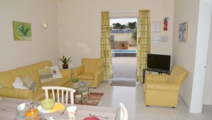 Sala de estar con sofás amarillos, una mesa de comedor con alimentos para el desayuno, un televisor y vistas a una piscina y un edificio.