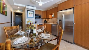 A modern kitchen with wooden cabinets, a stainless steel refrigerator, and a glass dining table set for four. - Mana Kai 404, Beautifully Renovated, Unbelievable Ocean View (Kihei)