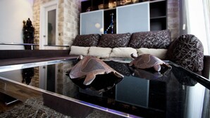 Een moderne woonkamer met een glazen salontafel, een accentmuur van steen, een bank met gestreepte kussens en een wandplank met decoratieve objecten.