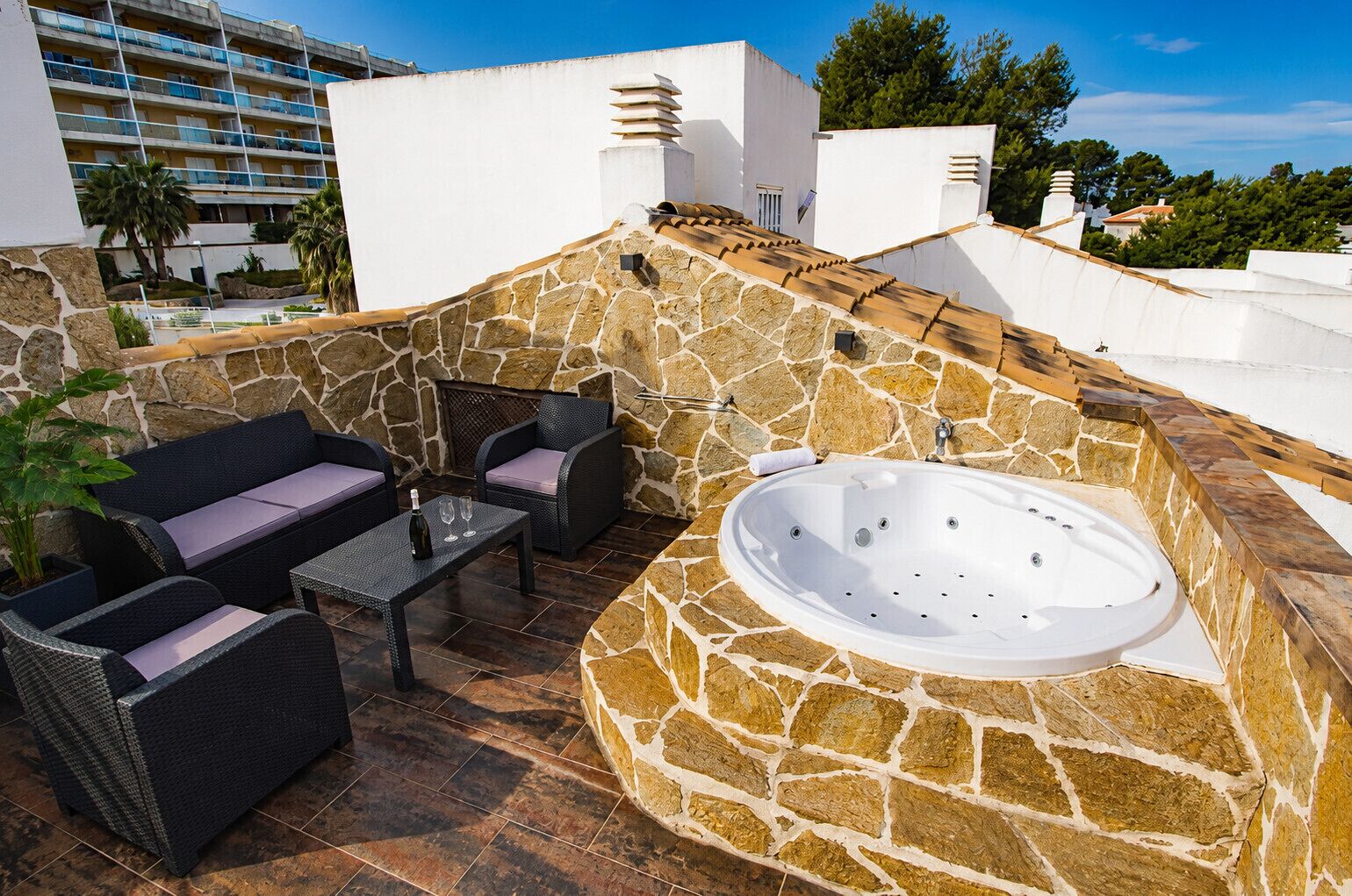 Une terrasse extérieure avec des murs en pierre, un jacuzzi, des meubles en osier et une vue sur un bâtiment en arrière-plan.