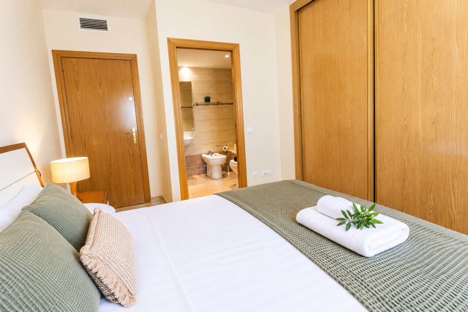 A hotel room with a bed, a bedside lamp, a green throw blanket, and a towel with a plant on it.