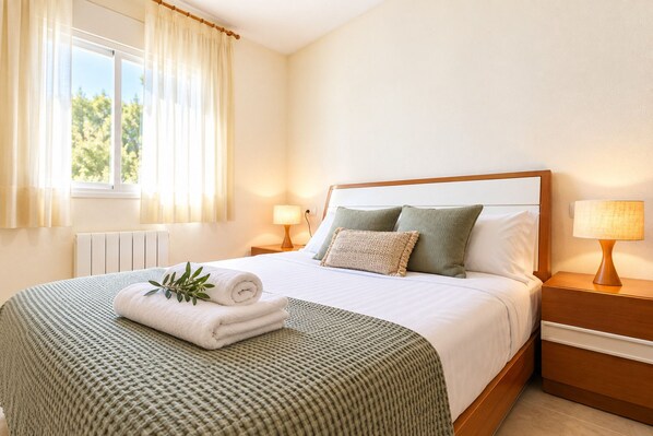 A neatly made bed with a rolled towel and a plant, a bedside lamp, and a window with curtains.