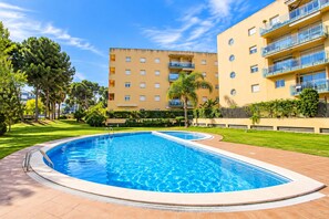 A swimming pool with a tiled deck, surrounded by well-maintained grass and trees, with a multi-story building in the background.
