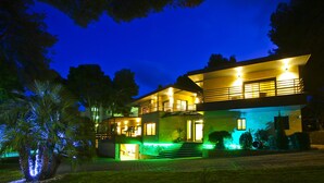 Een tweelaags huis met een balkon, verlicht met warme verlichting, omringd door groen.