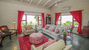 A spacious living room with a white sofa, red curtains, a round ottoman, and a ceiling fan.