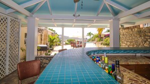 A covered outdoor bar area with a pool in the background, a ceiling fan, and a view of the pool and landscape.