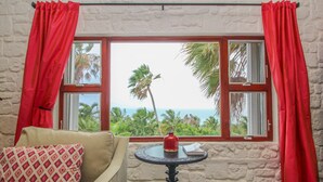 A room with a view of the ocean, a sofa, a small table with a vase, and red curtains.