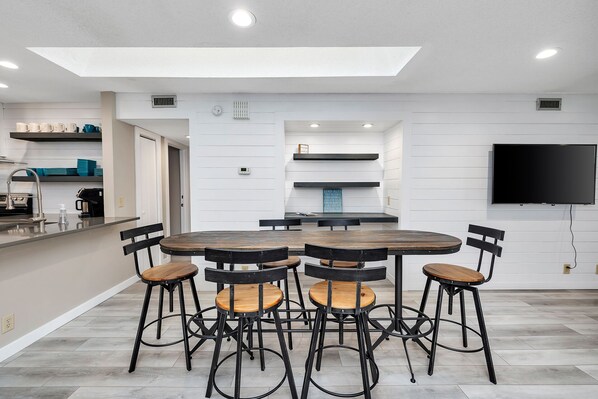 A modern kitchen with a round wooden table and black metal chairs, a built-in oven, and a flat-screen TV mounted on the wall.
