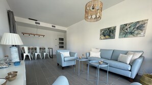 Un salon moderne avec un canapé bleu, deux tables d’appoint, un coin repas avec des chaises blanches et un espace cuisine.