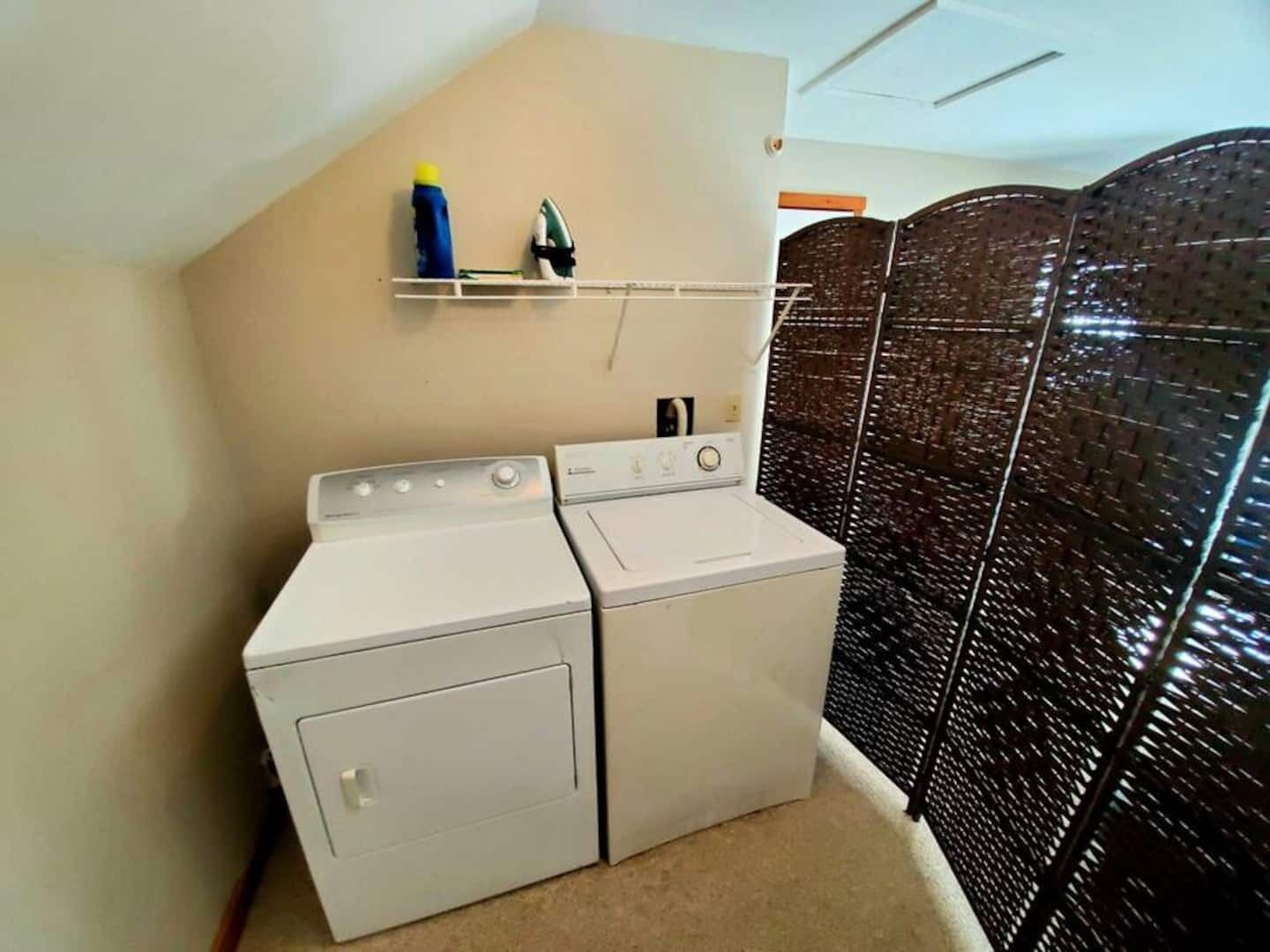 A laundry area with two white washing machines, a shelf with a bottle and an iron, and a dark patterned partition.