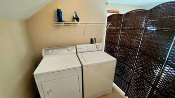 A laundry area with two white washing machines, a shelf with a bottle and an iron, and a dark patterned partition.