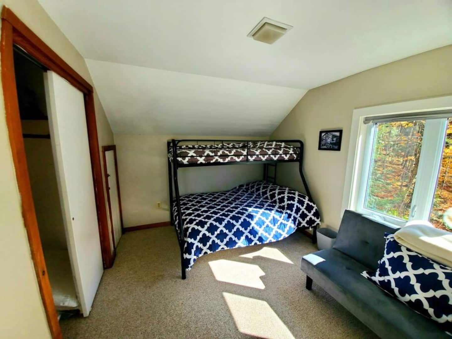 A room with a bunk bed, a sofa, a window with a view of trees, and a door leading to another room.