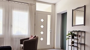 Ett modernt vardagsrum med en grå fåtölj, en svart pall och ett soffbord i glas. En vit dörr med fönster i form av romber syns, och den leder in till ett annat rum.