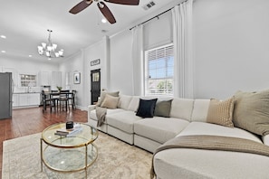 A spacious living room with a large sectional sofa, a glass coffee table, and a dining area with a kitchen in the background.