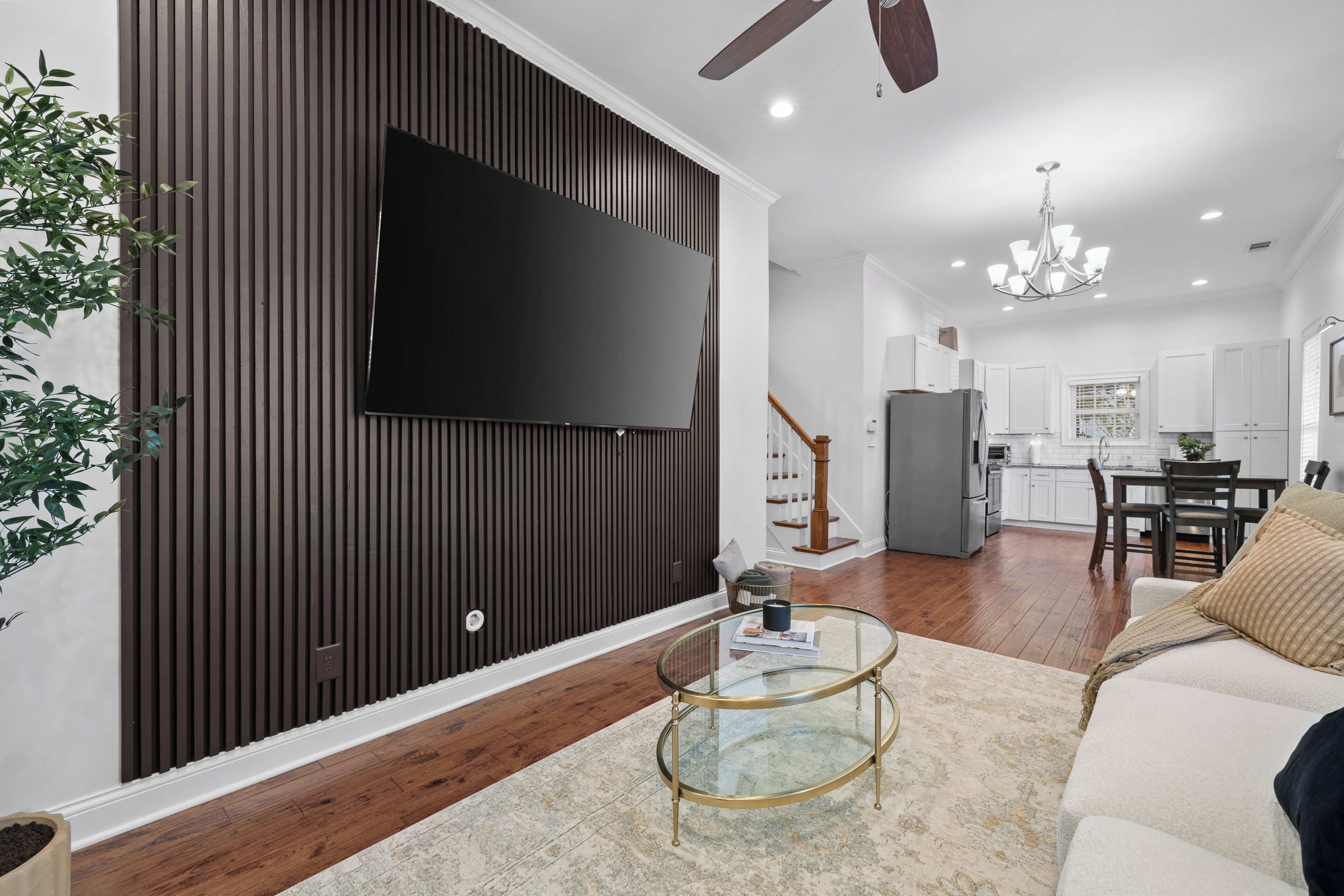 Un moderno salón con un televisor de pantalla plana montado en una pared oscura, una mesa de centro de cristal con una revista, un sofá blanco y una escalera que lleva a una zona de cocina.