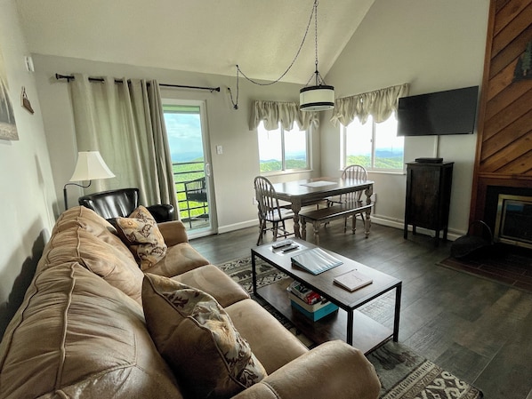A living room with a sofa, coffee table, and a dining area with chairs and a table. There is a TV on a stand and a fireplace.