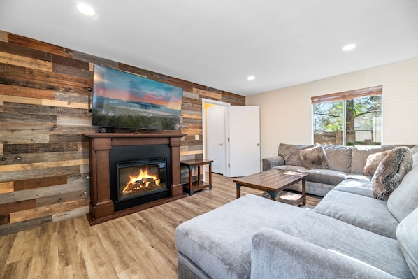 A living room with a fireplace, a large flat-screen TV mounted on a wooden wall, a grey sofa, and a wooden coffee table.