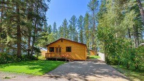 A wooden cabin with a porch, surrounded by tall trees and a gravel driveway.