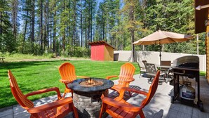 An outdoor patio with a stone fire pit table surrounded by orange Adirondack chairs, a barbecue grill, and a propane tank.