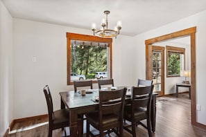 A dining area with a wooden table and chairs, a chandelier, and large windows with a view of trees and vehicles outside.