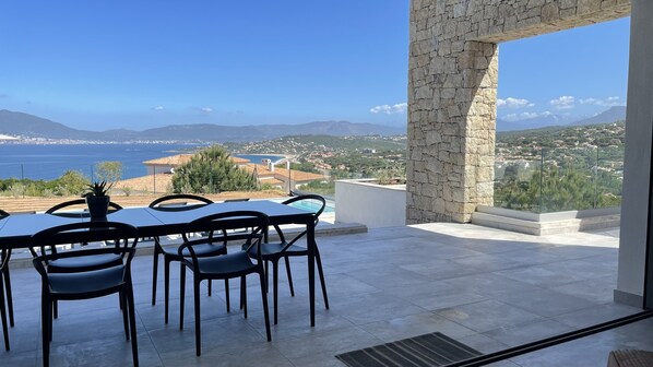Un espace repas comprenant une table et des chaises, un pilier en pierre, et une vue sur la mer et le paysage environnant.