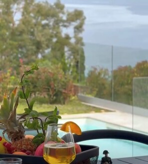 Un verre de bière avec une tranche de citron, posé sur une table où se trouvent des fruits et une petite figurine, donnant sur une piscine et une végétation luxuriante.