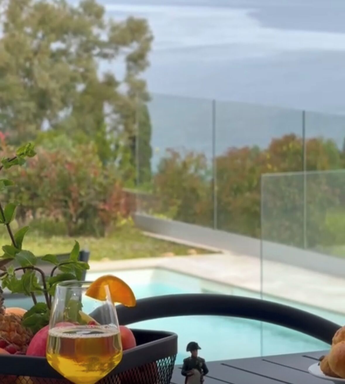 Un vaso de líquido amarillo, posiblemente una bebida, sobre una mesa con una cesta que contiene frutas y una pequeña figurina.