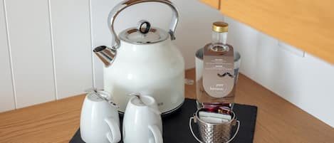 Una tetera blanca, dos tazas de té, una botella de licor y un vaso metálico sobre un posavasos negro.