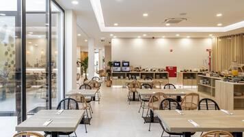 Un comedor amplio con varias mesas y sillas, una zona de cocina con electrodomésticos y estantes, y un muro de cristal que ofrece vistas al área de estar exterior.
