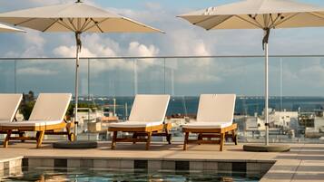 A rooftop pool area with lounge chairs and umbrellas, overlooking a marina and the sea.