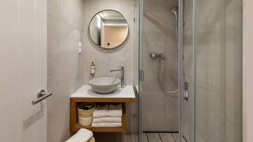 Un baño moderno con ducha envuelta en vidrio, espejo redondo, lavamanos sobre encimera y repisa de madera con toallas.
