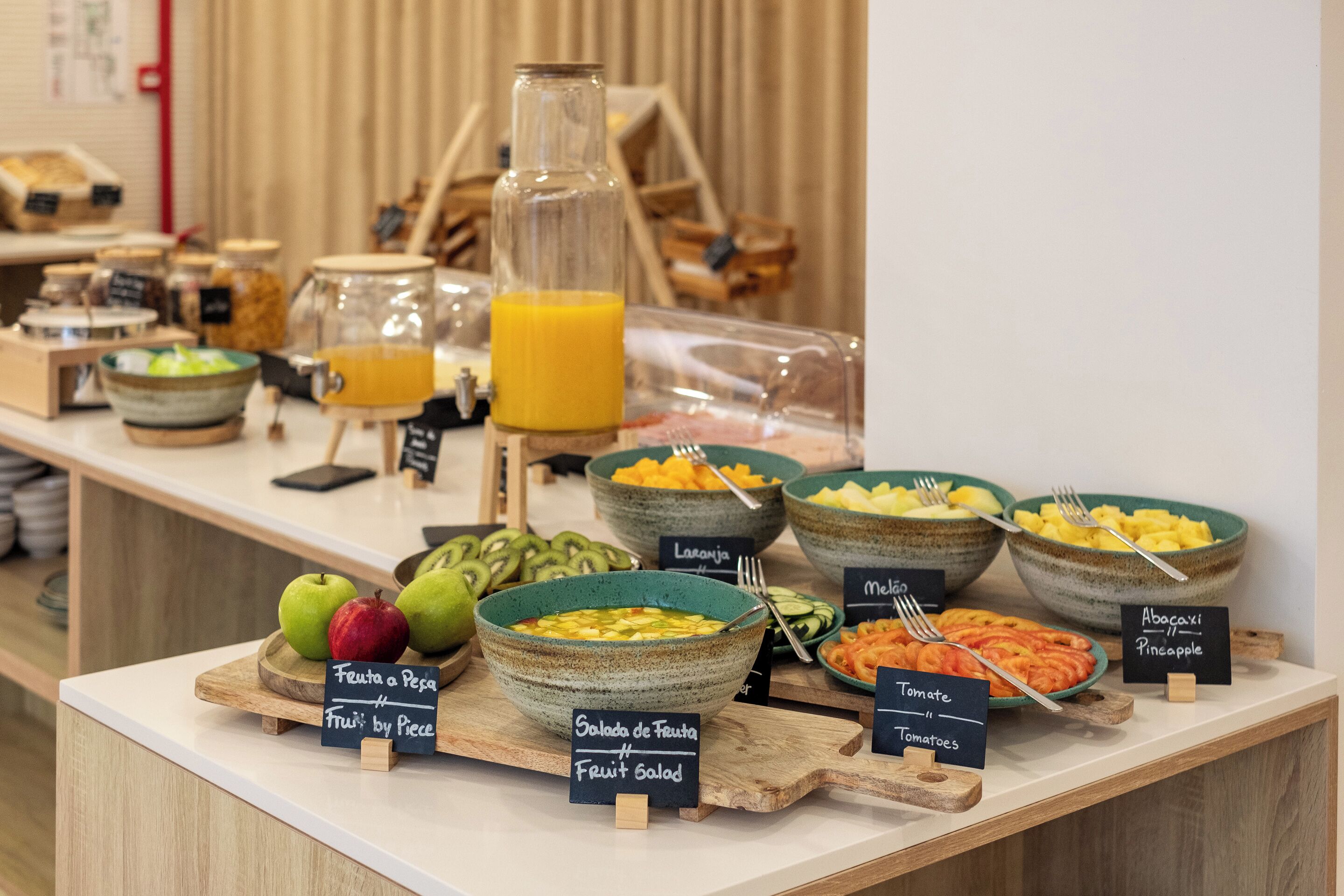 A breakfast buffet with various fruit bowls, a juice dispenser, and labeled signs indicating different fruit options.