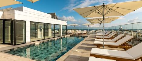 A rooftop infinity pool with sun loungers and umbrellas, surrounded by glass walls offering city views.