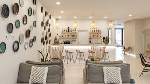 A modern living room with a kitchen in the background, featuring a bar area with stools and a variety of bottles. - Pure Formosa Concept Hotel (Olhao)