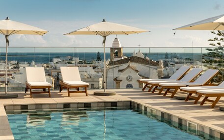 A rooftop pool area with lounge chairs and umbrellas, overlooking a coastal cityscape.