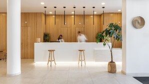 A modern reception area with a wooden panel wall, a reception desk, two wooden stools, and a large potted plant. - Pure Formosa Concept Hotel (Olhao)