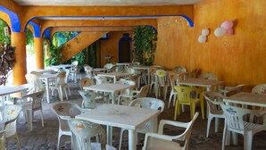 Uma área de refeições ao ar livre com mesas e cadeiras de plástico branco, cercada por paredes laranja e arcos azuis.