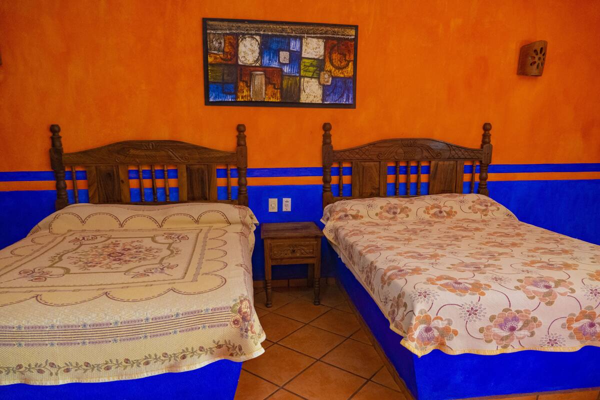 Dos camas de madera con ropa de cama floral, una mesita de noche de madera pequeña y un cuadro abstracto colorido en la pared naranja.