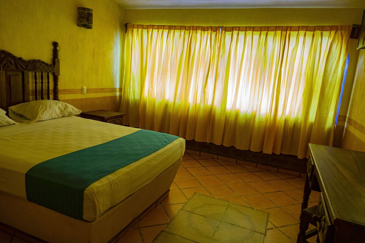 Una habitación de hotel con una cama grande, un cabecero de madera, una mesita de noche y un ventanal con cortinas amarillas.