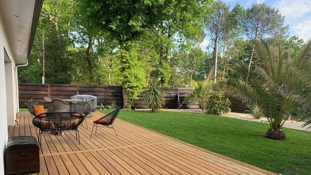 Une terrasse en bois avec des meubles d’extérieur, une table abritée et un jardin clos, agrémenté d’arbres et de plantes.