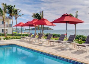A poolside area with lounge chairs and red umbrellas, overlooking a body of water. - Dawn Beach Club Resort Sint Maarten (Oyster Pond)